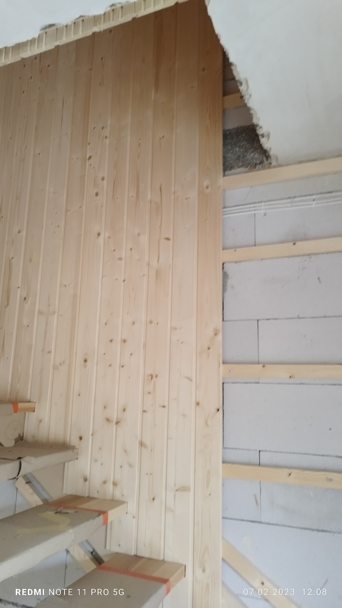 Montaż boazerii drewnianej na ścianie z bloczków betonowych, widoczne surowe schody i fragment uszkodzonego sufitu.