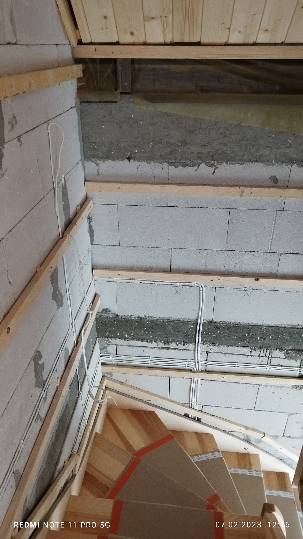 Wnętrze w trakcie prac wykończeniowych: surowa ściana z betonu komórkowego, drewniana konstrukcja pod sufitem, instalacja elektryczna w peszlach oraz fragment drewnianych schodów z zabezpieczeniami...