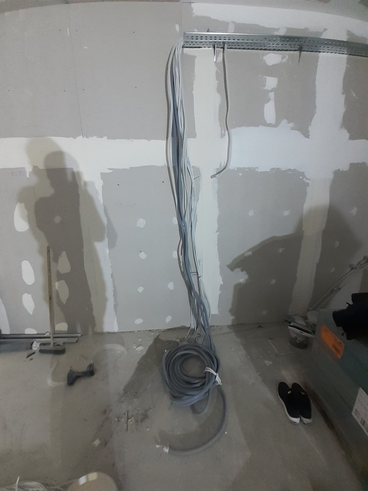 Wnętrze w trakcie prac wykończeniowych: ściany z płyt kartonowo-gipsowych, widoczne przewody elektryczne zwisające z metalowej szyny, narzędzia i buty robocze na podłodze.