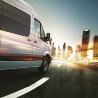 Biały bus transportowy w ruchu na tle panoramy miasta z rozmytymi wieżowcami, widok z boku, dynamiczne ujęcie.