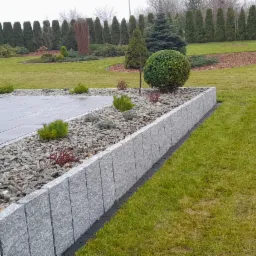 Nowoczesny ogród z szarą kostką brukową, kamiennym obrzeżem rabaty z drobnymi kamykami i niskimi roślinami, równo skoszonym trawnikiem i rzędem wysokich, zielonych drzew w tle.