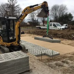 Żółta mini koparka CAT podnosi betonowy element za pomocą zawiesia linowego, przygotowany teren z piaskiem, ubijarka w tle, paleta z betonowymi płytami z otworami po lewej stronie.