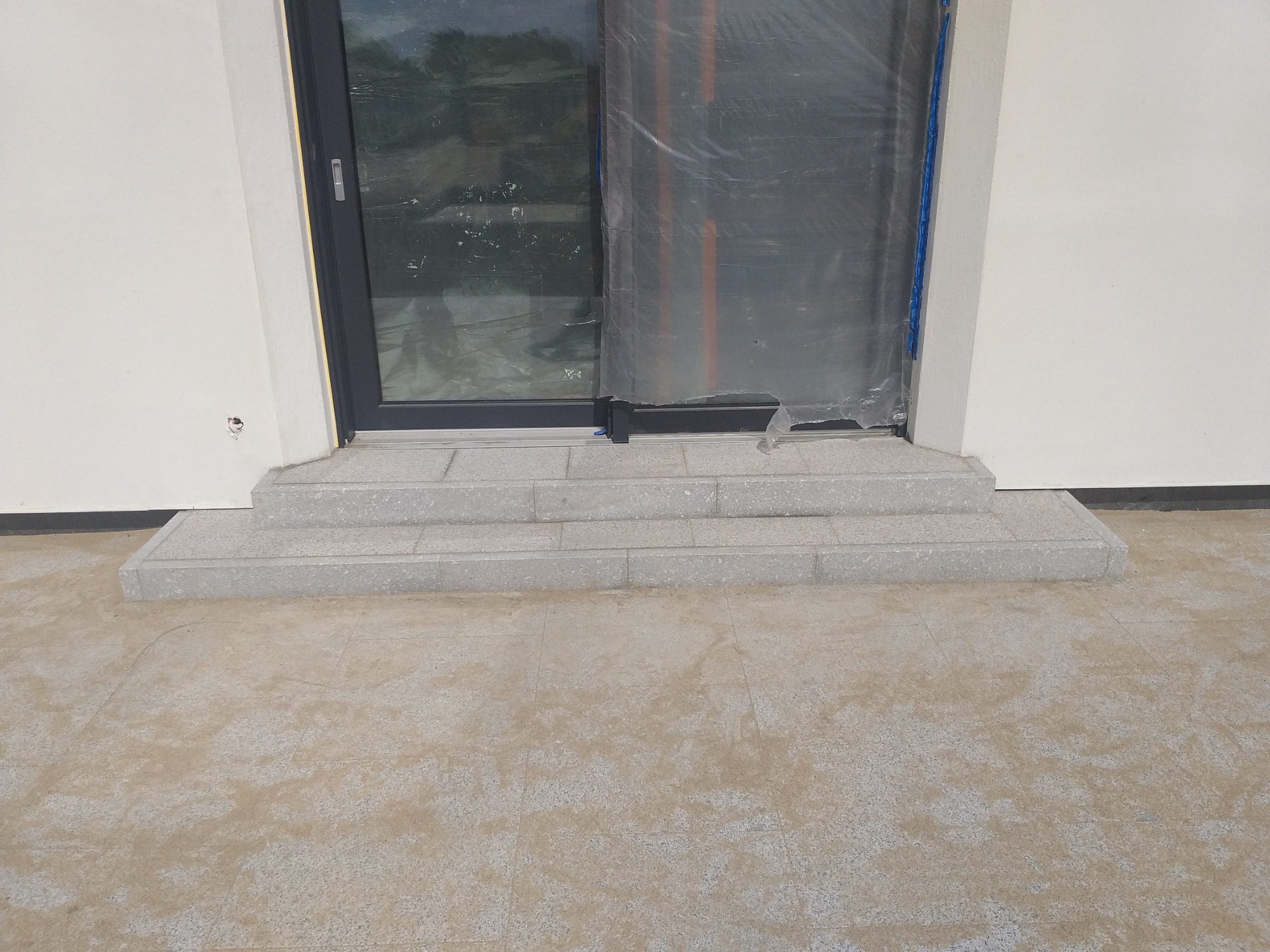 Schody wejściowe wyłożone jasnoszarą kostką brukową prowadzące do drzwi tarasowych zabezpieczonych folią ochronną, widoczne płytki chodnikowe.