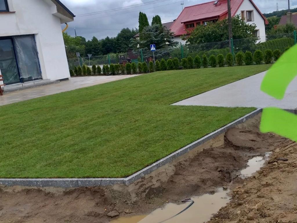 Świeżo założony trawnik z rolki przy nowoczesnym domu, widoczne krawężniki i przygotowane podłoże w wykopie, fragment chodnika z kostki, pochmurne niebo.