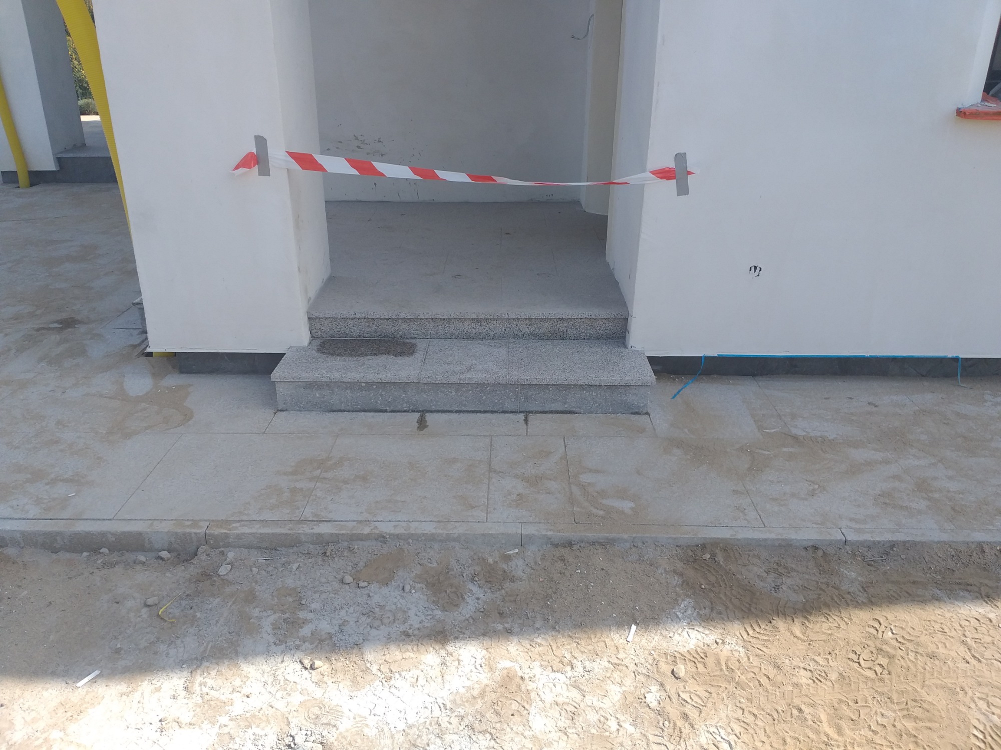 Ułożona szara kostka brukowa przed wejściem do budynku, zabezpieczona taśmą ostrzegawczą, widoczne ślady prac ziemnych.