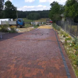 Ułożona, brązowo-czerwona kostka brukowa na podjeździe, z widocznymi elementami drenażu, obrzeżami z ciemnej kostki, roślinnością po bokach, ciężarówką i koparką w tle.