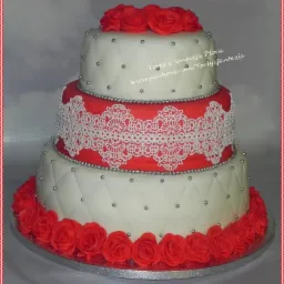 Trzypiętrowy tort weselny w kolorach białym i czerwonym, zdobiony wzorem rombów z srebrnymi koralikami i koronkową aplikacją, wykończony czerwonymi różami.