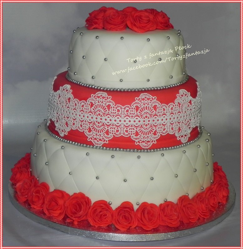 Trzypiętrowy tort weselny w kolorach białym i czerwonym, zdobiony wzorem rombów z srebrnymi koralikami i koronkową aplikacją, wykończony czerwonymi różami.