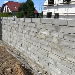 Wznoszenie szarego muru z bloczków betonowych w Dąbcze, widoczna poziomica i fragment gruntu, w tle dom w budowie ze rusztowaniem.