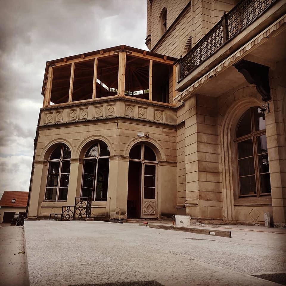 Budynek w trakcie renowacji z dobudowywaną drewnianą konstrukcją balkonu, widoczne okna z łukami, poziomica i wiadro na posadzce.