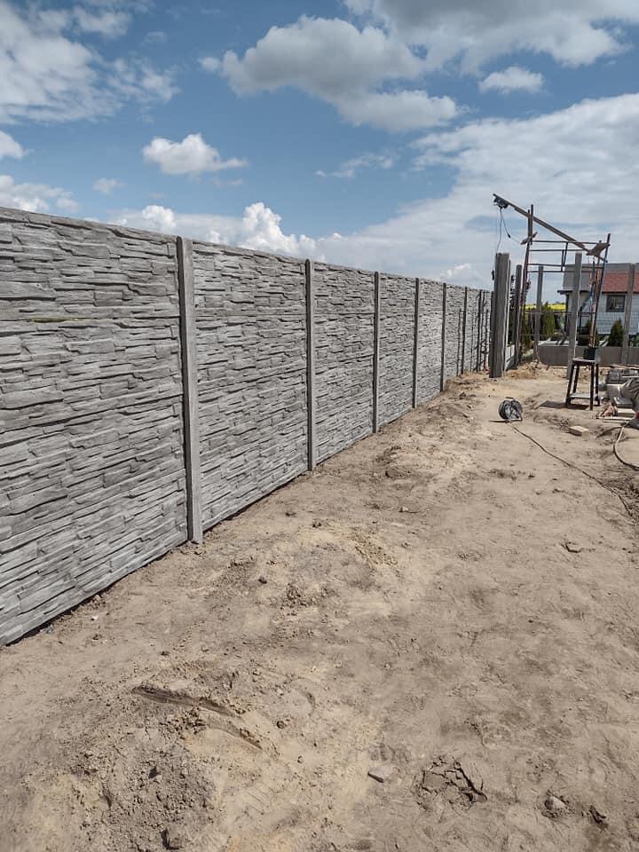 Długi, szary, betonowy płot z elementami imitującymi kamień, w tle błękitne niebo z obłokami i częściowo widoczny dom w trakcie budowy.