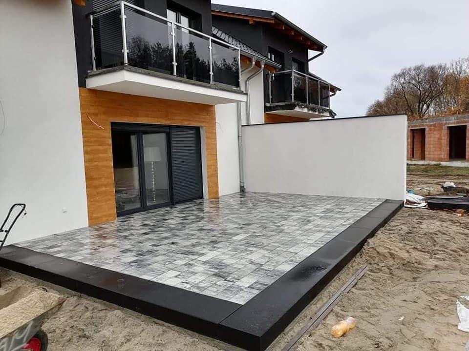 Nowoczesny taras z szaro-białej kostki brukowej, obramowany czarnym obrzeżem, przylegający do budynku z balkonami i elementami drewnopodobnymi, w tle surowy budynek w trakcie budowy.