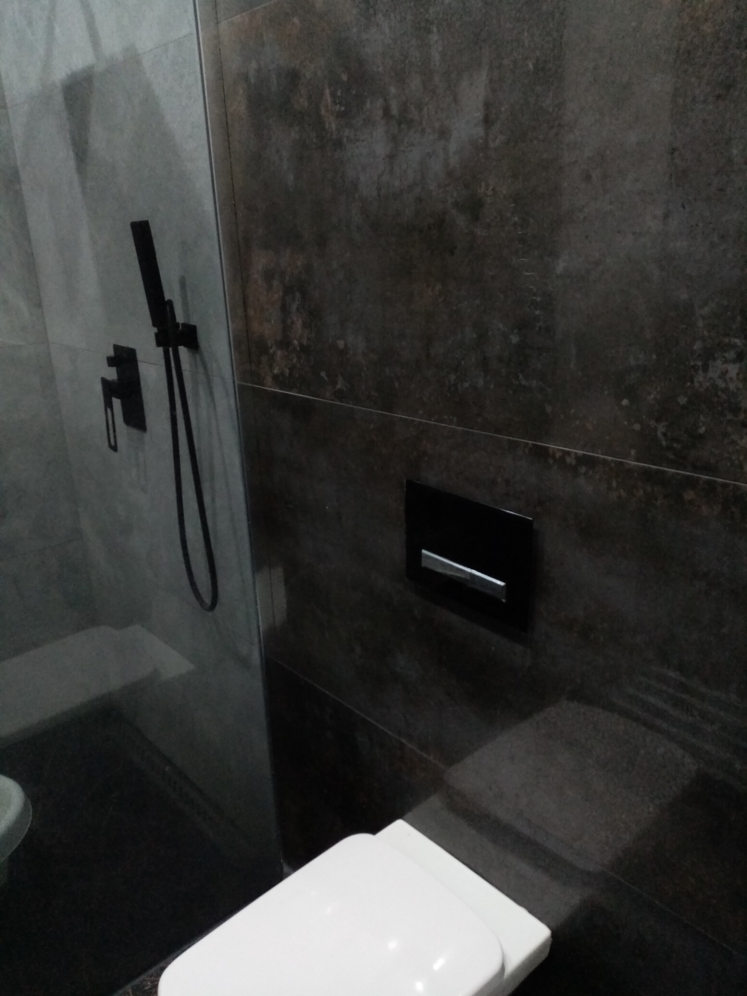 Nowoczesna łazienka z ciemnymi płytkami imitującymi kamień, czarną armaturą prysznicową za szklaną ścianką i wiszącą miską WC z czarnym przyciskiem spłukującym.