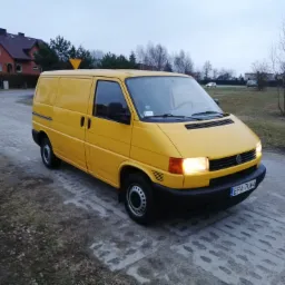 Żółty van Volkswagen Transporter T4 zaparkowany na betonowej drodze, z włączonymi światłami mijania, na tle domów i drzew.