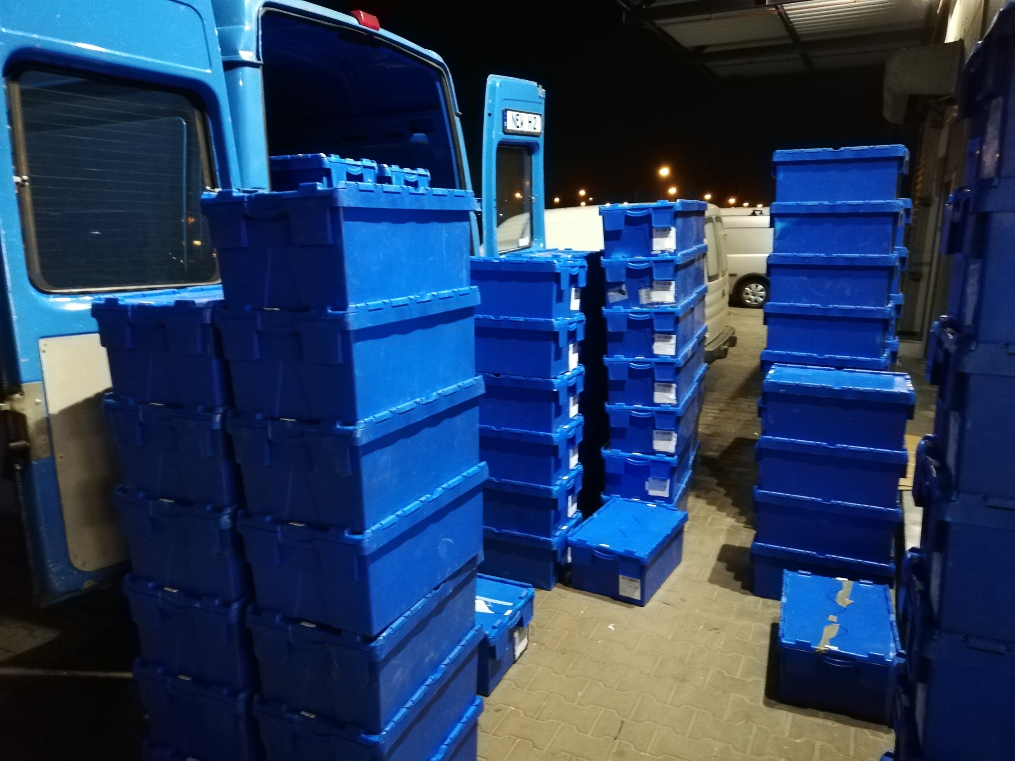 Stosy niebieskich, plastikowych pojemników ustawione na chodniku przy otwartym furgonie w nocnym oświetleniu, gotowe do załadunku lub rozładunku.