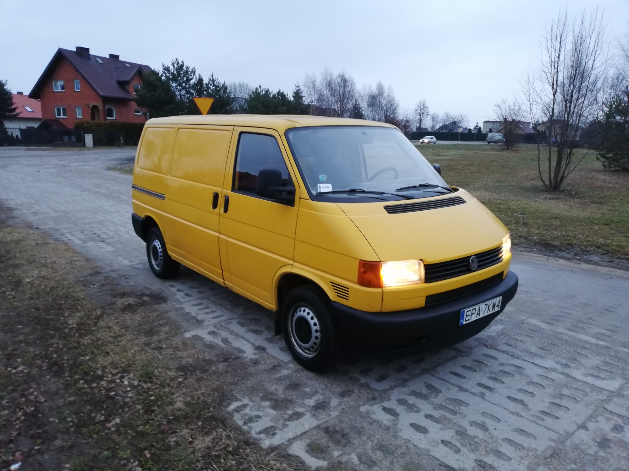 Żółty van Volkswagen Transporter T4 zaparkowany na betonowej drodze, z włączonymi światłami mijania, na tle domów i drzew.