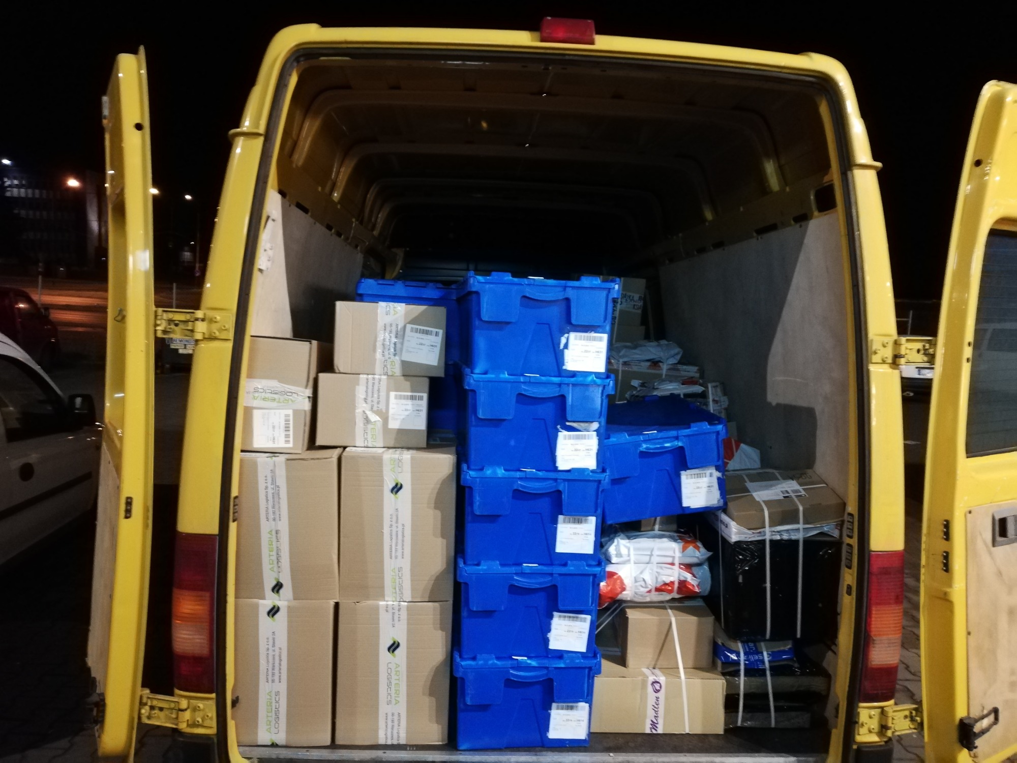 Wnętrze żółtego busa załadowane kartonami z logo Arteria Logistics i niebieskimi plastikowymi pojemnikami, widok z tyłu, otwarte drzwi.