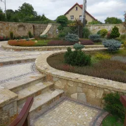 Kaskadowy ogród z kamiennymi murkami oporowymi i ścieżką z kostki brukowej, z różnorodną roślinnością ozdobną i drzewami iglastymi, na tle domu jednorodzinnego.