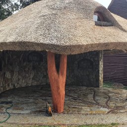 GP - MUR - Altana z kamienia i strzechy, z drewnianymi słupami imitującymi pnie drzew, czyszczona myjką ciśnieniową przez dwóch mężczyzn.