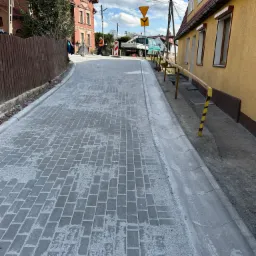 Świeżo ułożona, szara kostka brukowa na nowej drodze, widok z perspektywy ulicy, z widocznym znakiem drogowym i fragmentem pracujących osób w tle.