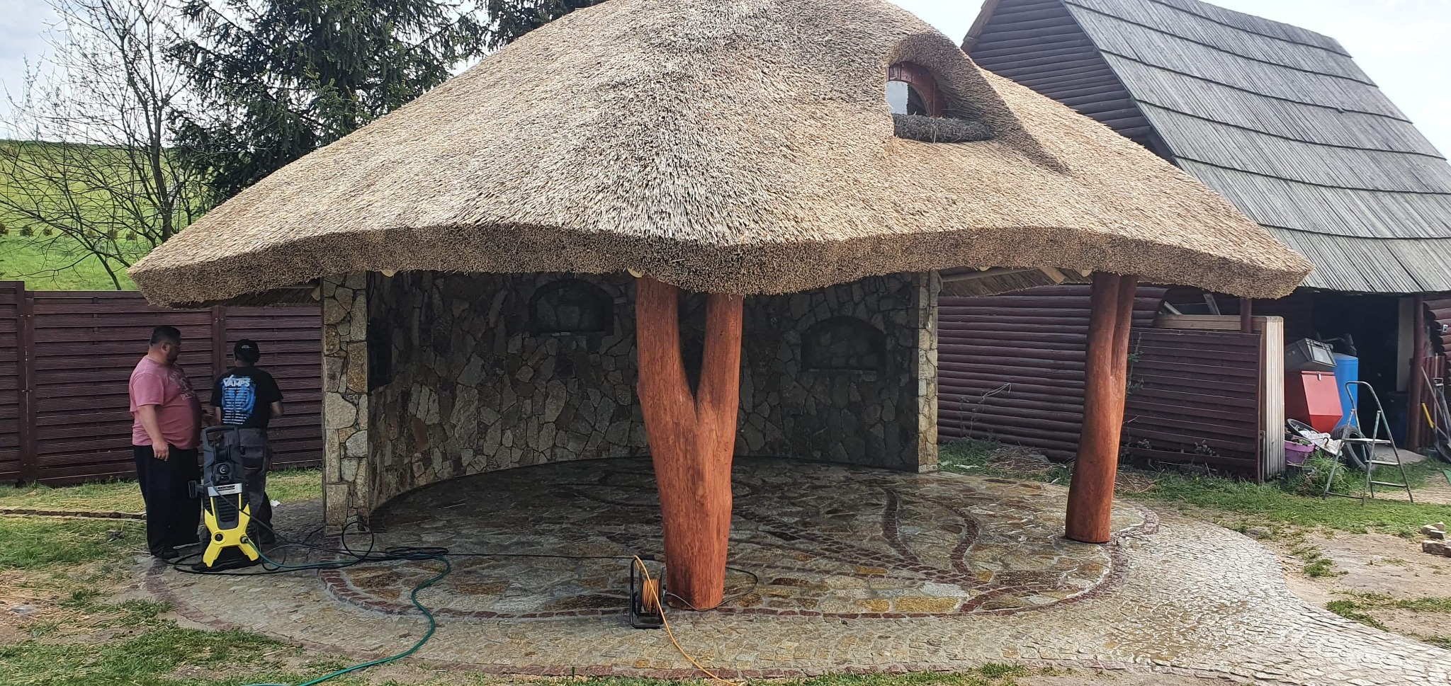 Altana z kamienia i strzechy, z drewnianymi słupami imitującymi pnie drzew, czyszczona myjką ciśnieniową przez dwóch mężczyzn.