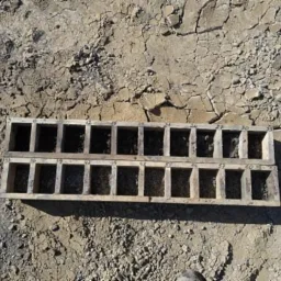 Drewniana forma z numerowanymi przegródkami wypełnionymi próbkami gruntu na wyschniętej, spękanej ziemi.