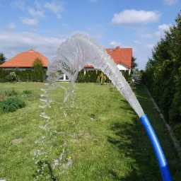 Strumień wody z niebieskiego węża, skierowany w górę, na tle zielonego trawnika, domu z pomarańczowym dachem i błękitnego nieba z chmurami.