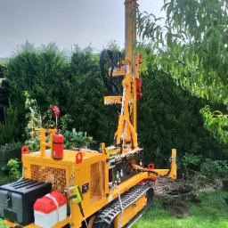 Żółta, gąsienicowa wiertnica do studni, z widocznym odwiertem w trawniku, na tle zielonych krzewów i drzew.