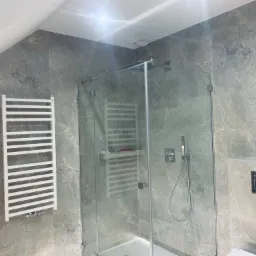 Nowoczesna łazienka z kabiną prysznicową ze szkła hartowanego, grzejnikiem drabinkowym na ścianie z płytek imitujących marmur oraz minimalistyczną armaturą.