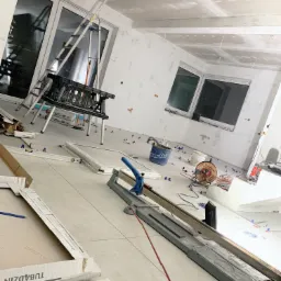 Wnętrze w trakcie prac wykończeniowych: położone płytki podłogowe, częściowo wykończony sufit podwieszany z płyt karton-gips, widoczna drabina, przecinarka do płytek, poziomica i inne narzędzia.