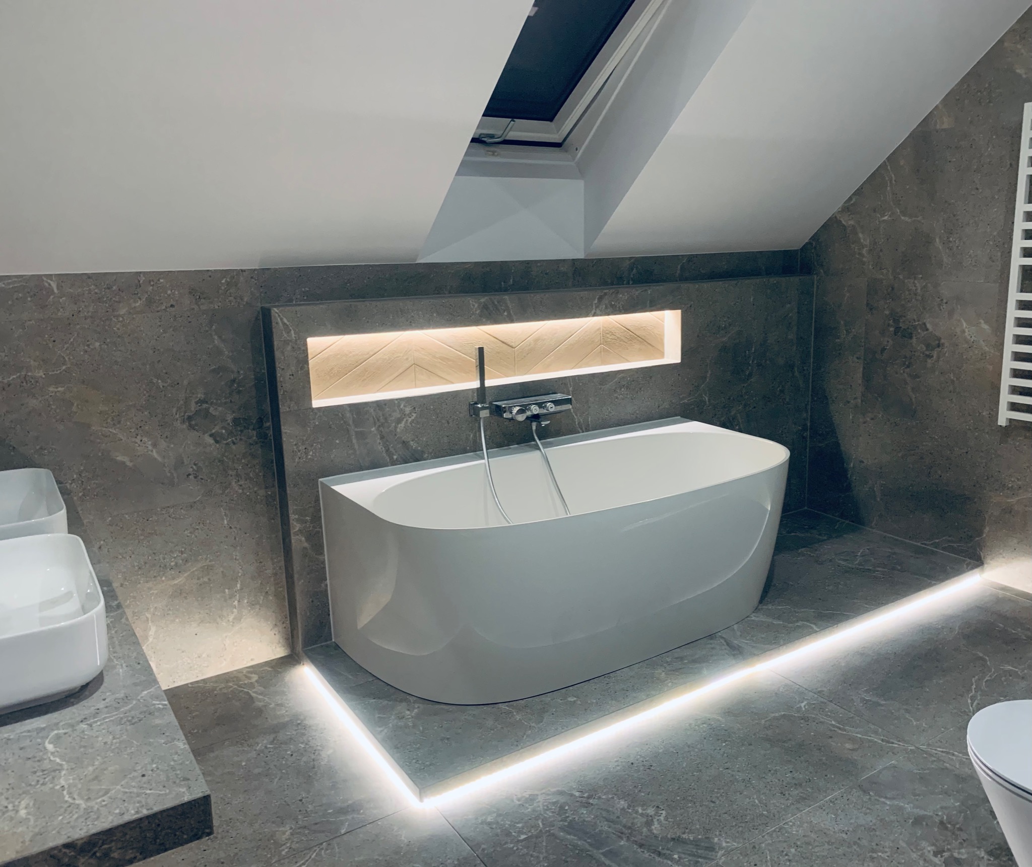 Nowoczesna łazienka na poddaszu z wolnostojącą białą wanną, szarą marmurową okładziną ścian i podłogi, oświetleniem LED wzdłuż podłogi i w niszy nad wanną z dekoracyjnym panelem drewnianym ułożonym...