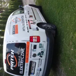 Biały samochód dostawczy z reklamą firmy Piątek Instalacje Elektryczne na tle zielonej trawy i drzew.