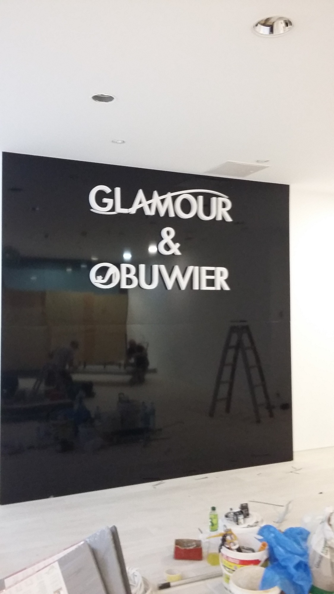 Czarna ściana z białym napisem 'GLAMOUR & OBUWIE', widoczne narzędzia i drabina, odblaski na ścianie, sugerujące montaż.