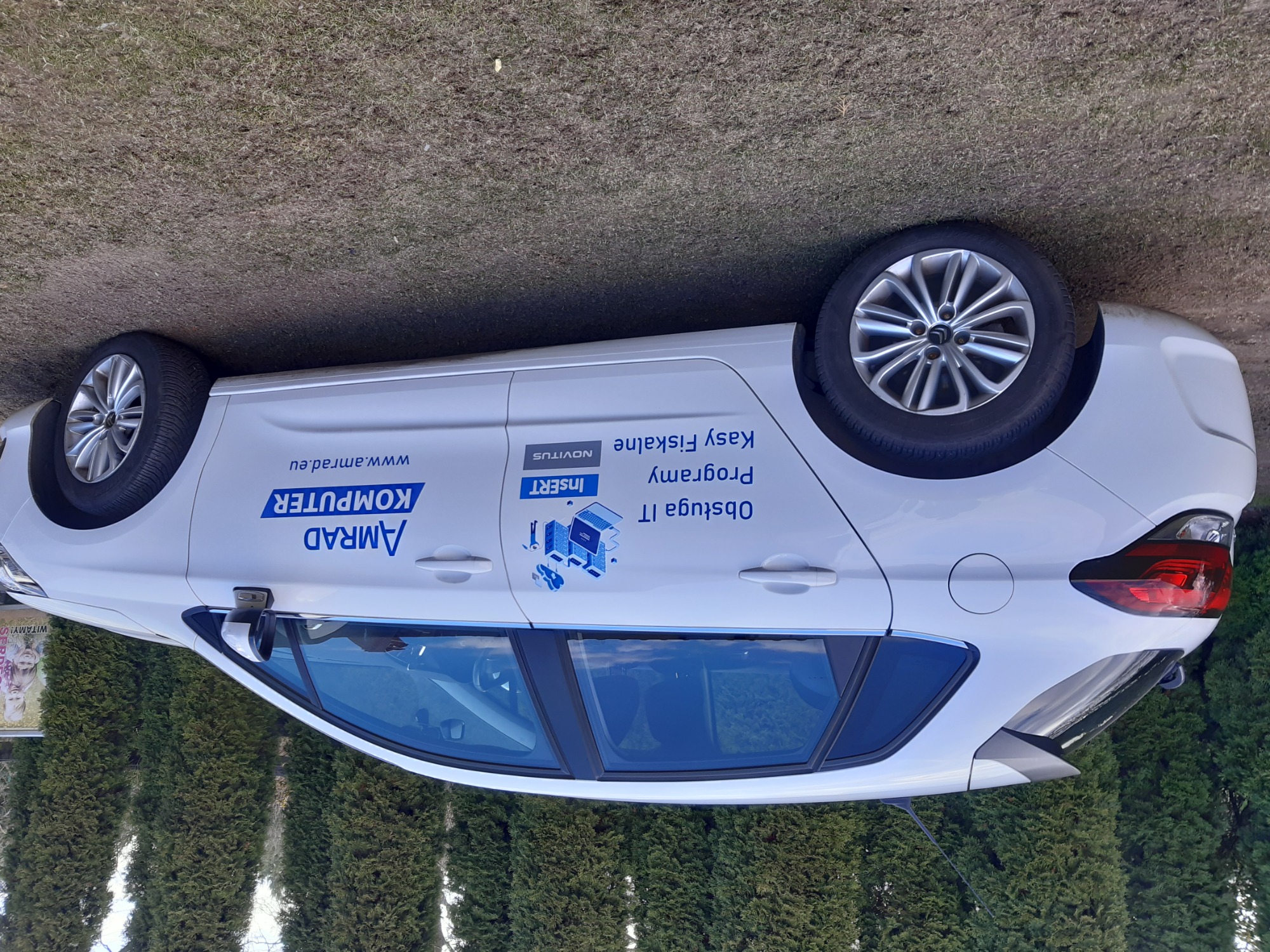 Biały samochód z reklamą firmy komputerowej AMRAD i usług IT, z widocznym logo i adresem strony internetowej, zaparkowany na tle zielonych krzewów.