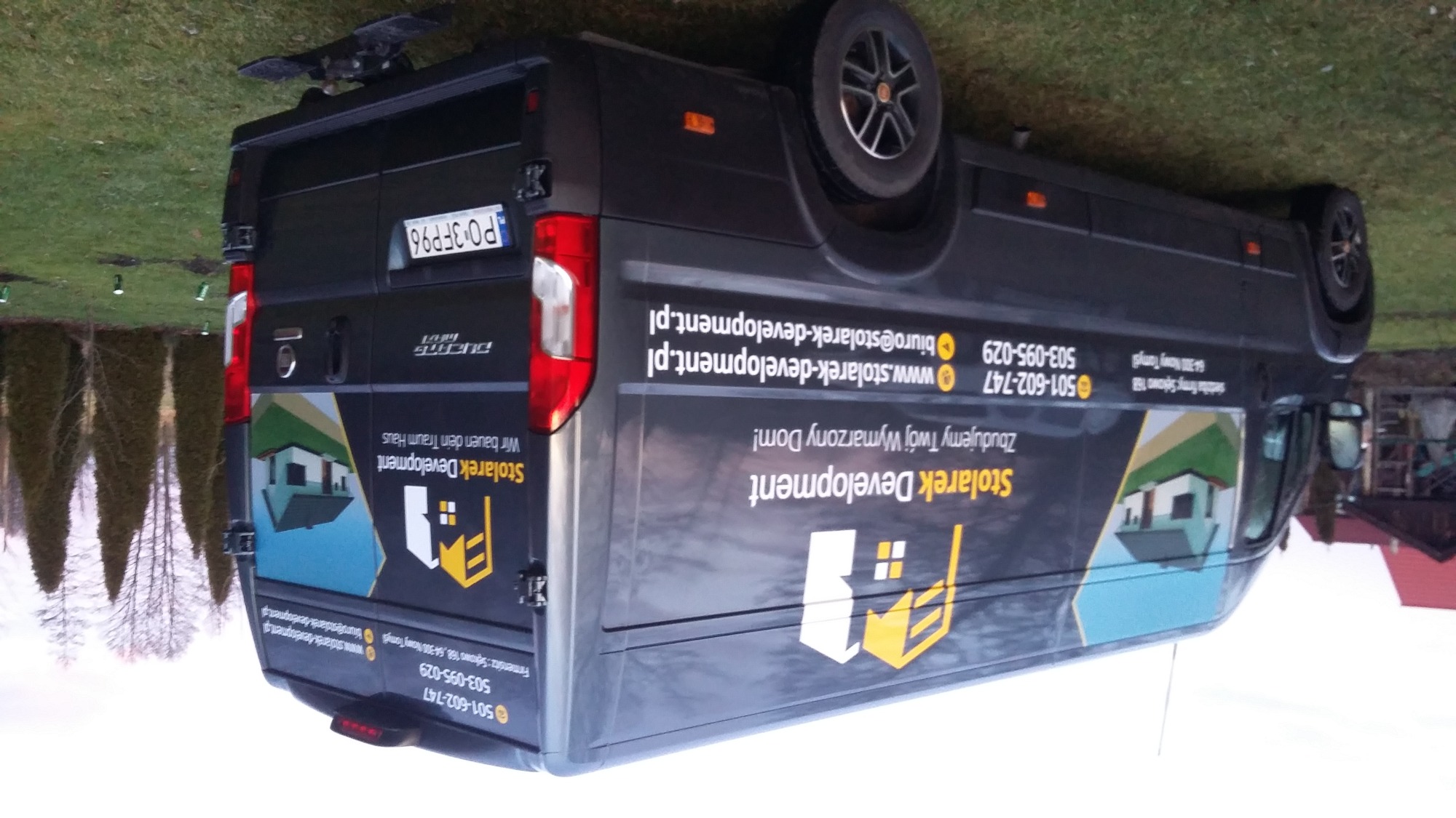 Czarny samochód dostawczy z reklamą firmy budowlanej Stolarek Development, zaparkowany na trawniku, widoczny numer rejestracyjny i dane kontaktowe.
