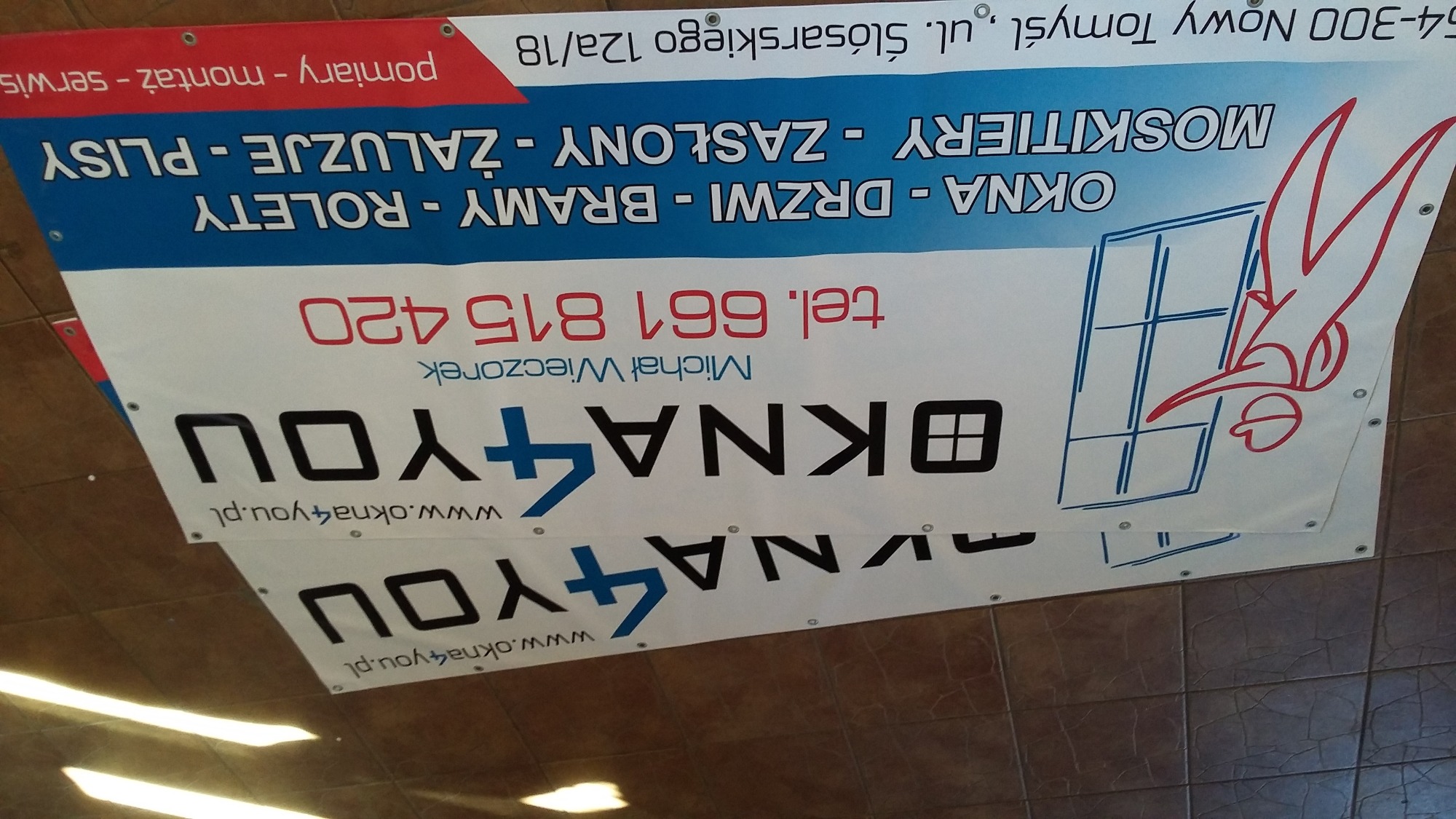 Dwa identyczne bannery reklamowe firmy oferującej okna, drzwi, bramy, rolety, moskitiery, zasłony, żaluzje i plisy, z widocznym logo, adresem strony internetowej i numerem telefonu, zawieszone...