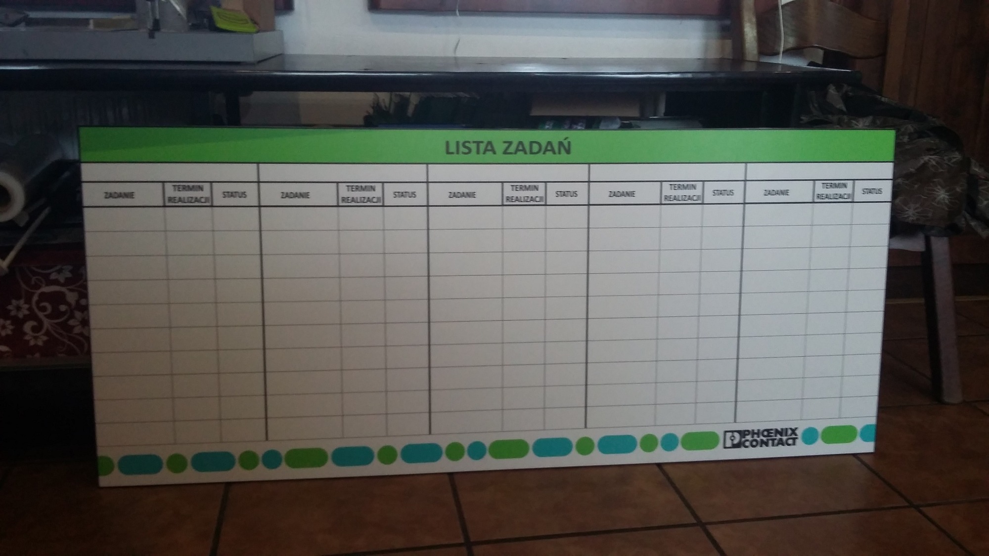 Tablica suchościeralna z nadrukowaną listą zadań do wypełnienia, z logo Phoenix Contact w prawym dolnym rogu, umieszczona na brązowych płytkach podłogowych