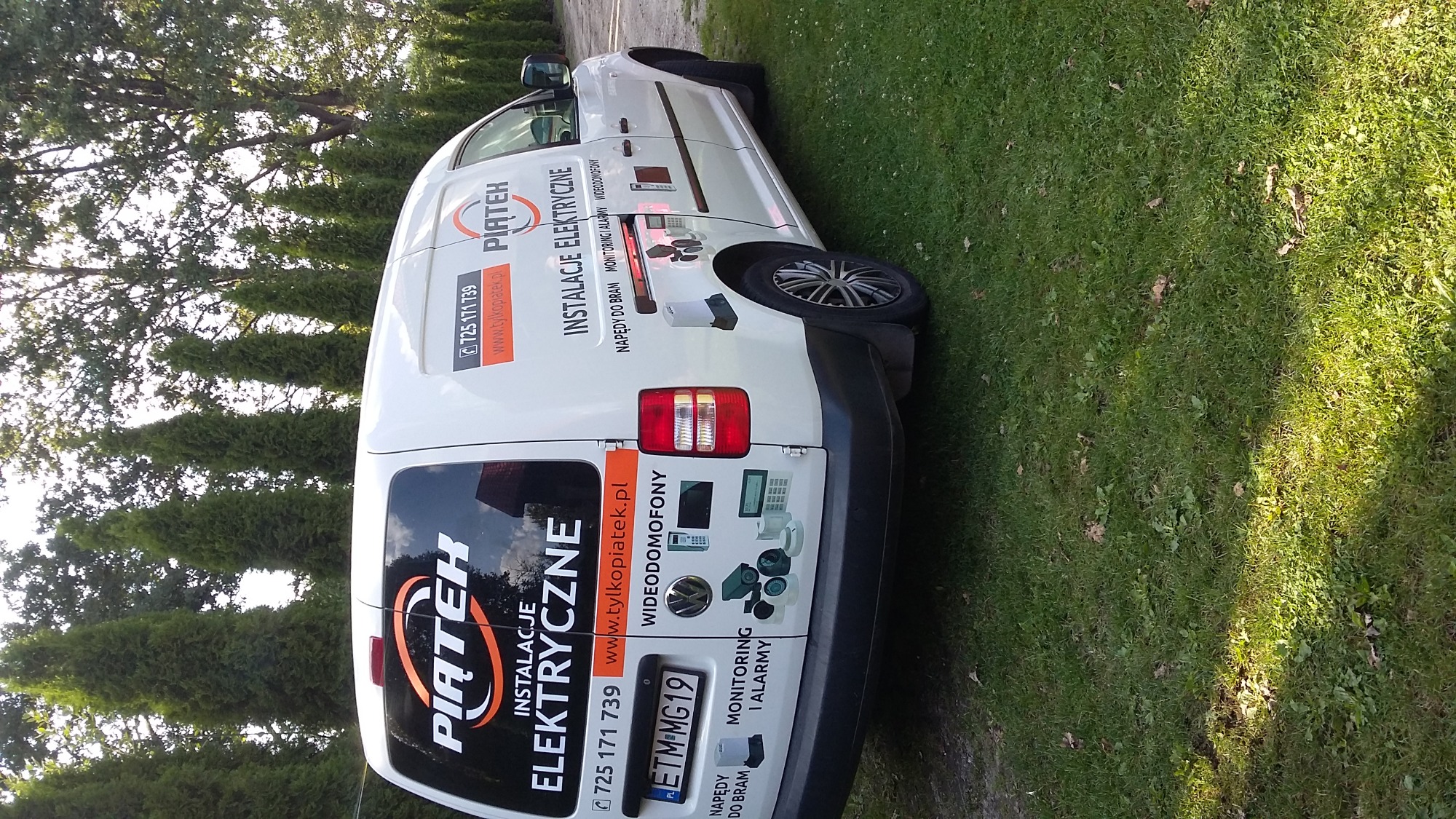 Biały samochód dostawczy z reklamą firmy Piątek Instalacje Elektryczne na tle zielonej trawy i drzew.