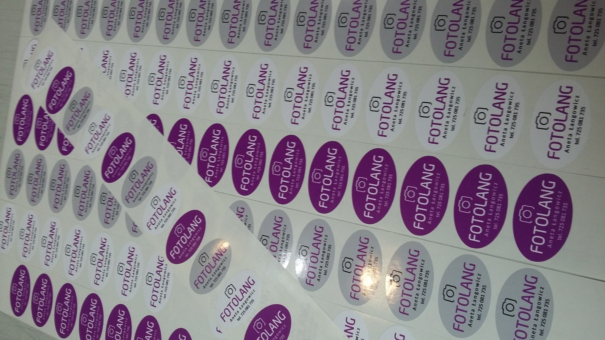 Arkusze z owalnymi, samoprzylepnymi naklejkami z logo 'FOTOLANG' i danymi kontaktowymi, częściowo odklejone od podłoża.