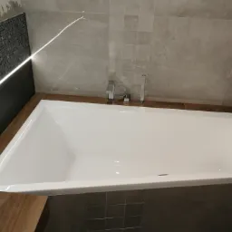 Nowoczesna, biała wanna w zabudowie z szarymi płytkami i mozaiką, bateria wannowa chromowana, oświetlenie LED wzdłuż ściany z mozaiką.