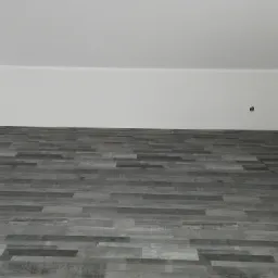 Ułożone panele podłogowe w odcieniach szarości, widok z dołu na białą ścianę i sufit z wystającym gniazdkiem elektrycznym.