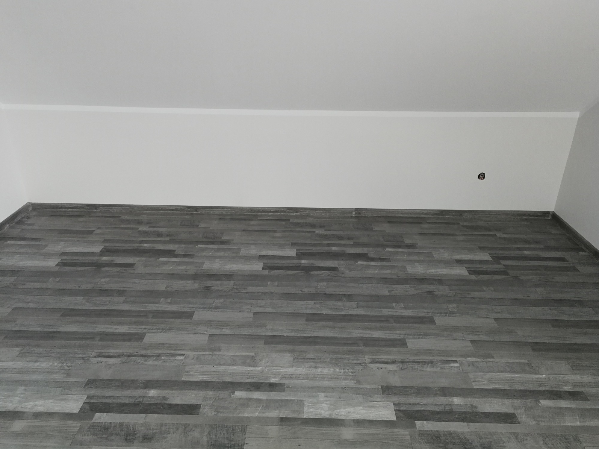 Ułożone panele podłogowe w odcieniach szarości, widok z dołu na białą ścianę i sufit z wystającym gniazdkiem elektrycznym.