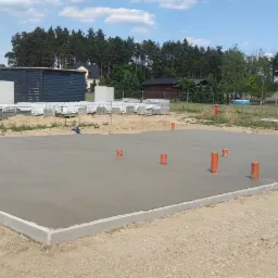 Wylana płyta fundamentowa z wystającymi pionami instalacyjnymi w Lubomyślu koło Żar, widok na plac budowy z częściowo wykończonym domem w tle.