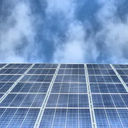 Rząd paneli słonecznych pod lekkim kątem, odbijających błękitne niebo z białymi chmurami.