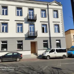 Odnowiona elewacja trzypiętrowej kamienicy w Rawa Mazowiecka z nowymi oknami i balkonami z kutą balustradą, widok z poziomu ulicy z zaparkowanymi samochodami.