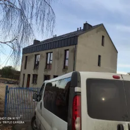 Surowy dom jednorodzinny z ciemnym, metalowym dachem, widoczny zza białego, lekko zabrudzonego samochodu dostawczego na tle placu budowy i błękitnego nieba.