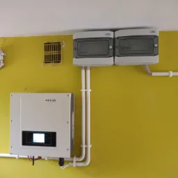 Zamontowany inwerter fotowoltaiczny Sofar z podłączonymi przewodami i dwoma rozdzielnicami na żółtej ścianie wewnątrz budynku.