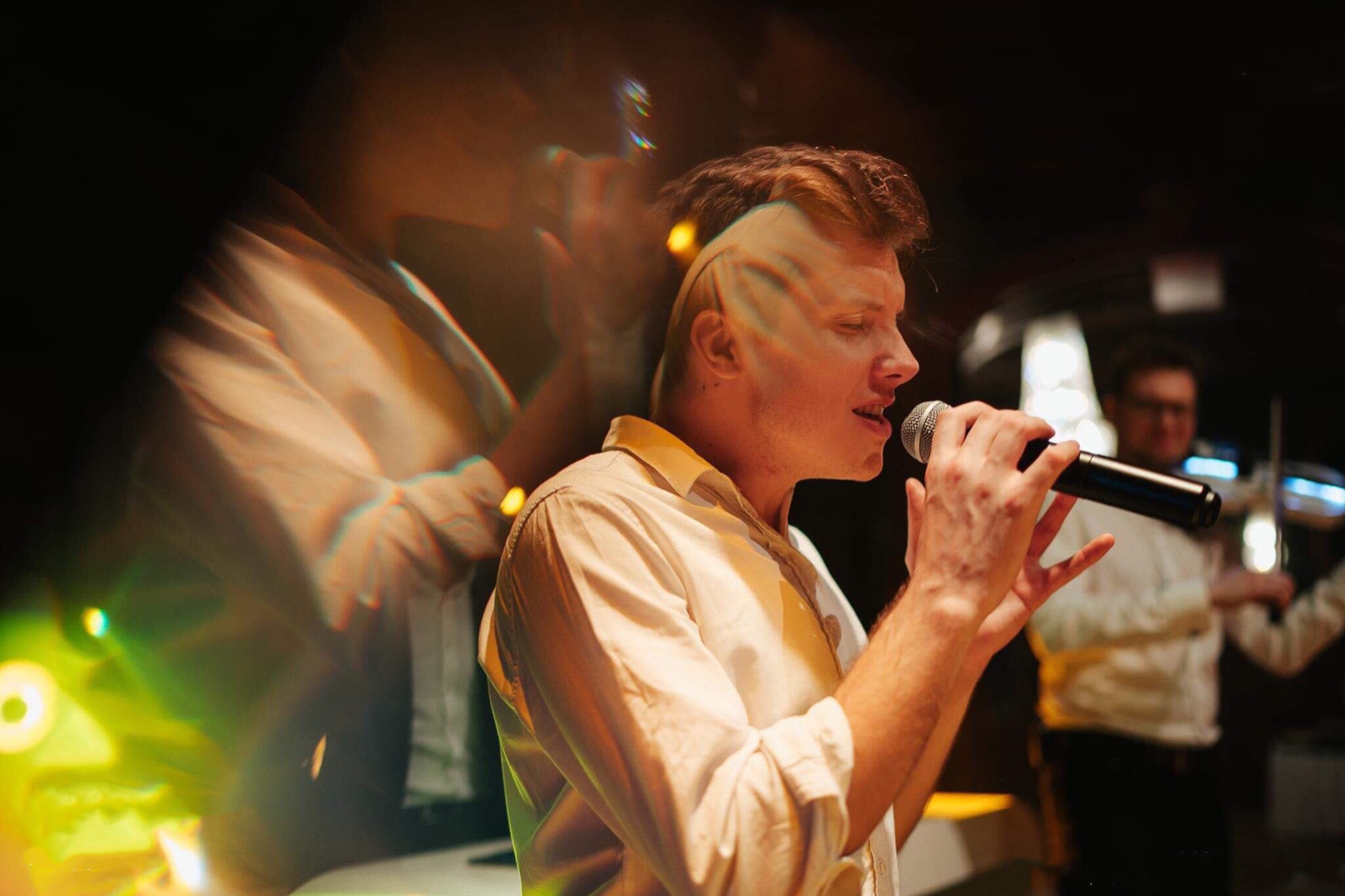 Wokalista z mikrofonem śpiewa na scenie, w tle muzyk gra na skrzypcach, efekt rozmycia i refleksów świetlnych.