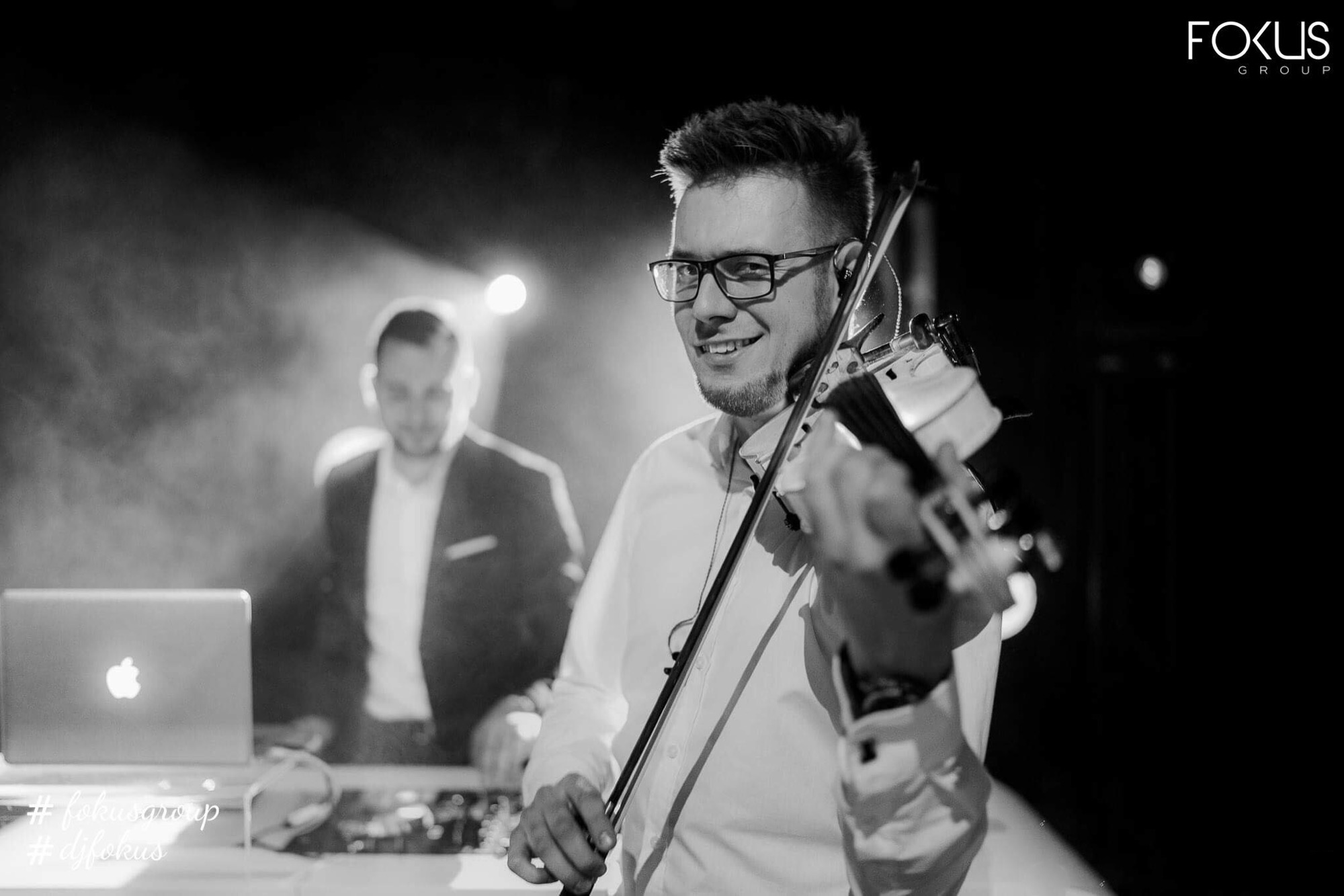 Uśmiechnięty skrzypek w okularach gra na skrzypcach na scenie, w tle laptop i drugi muzyk w marynarce, czarno-białe zdjęcie.