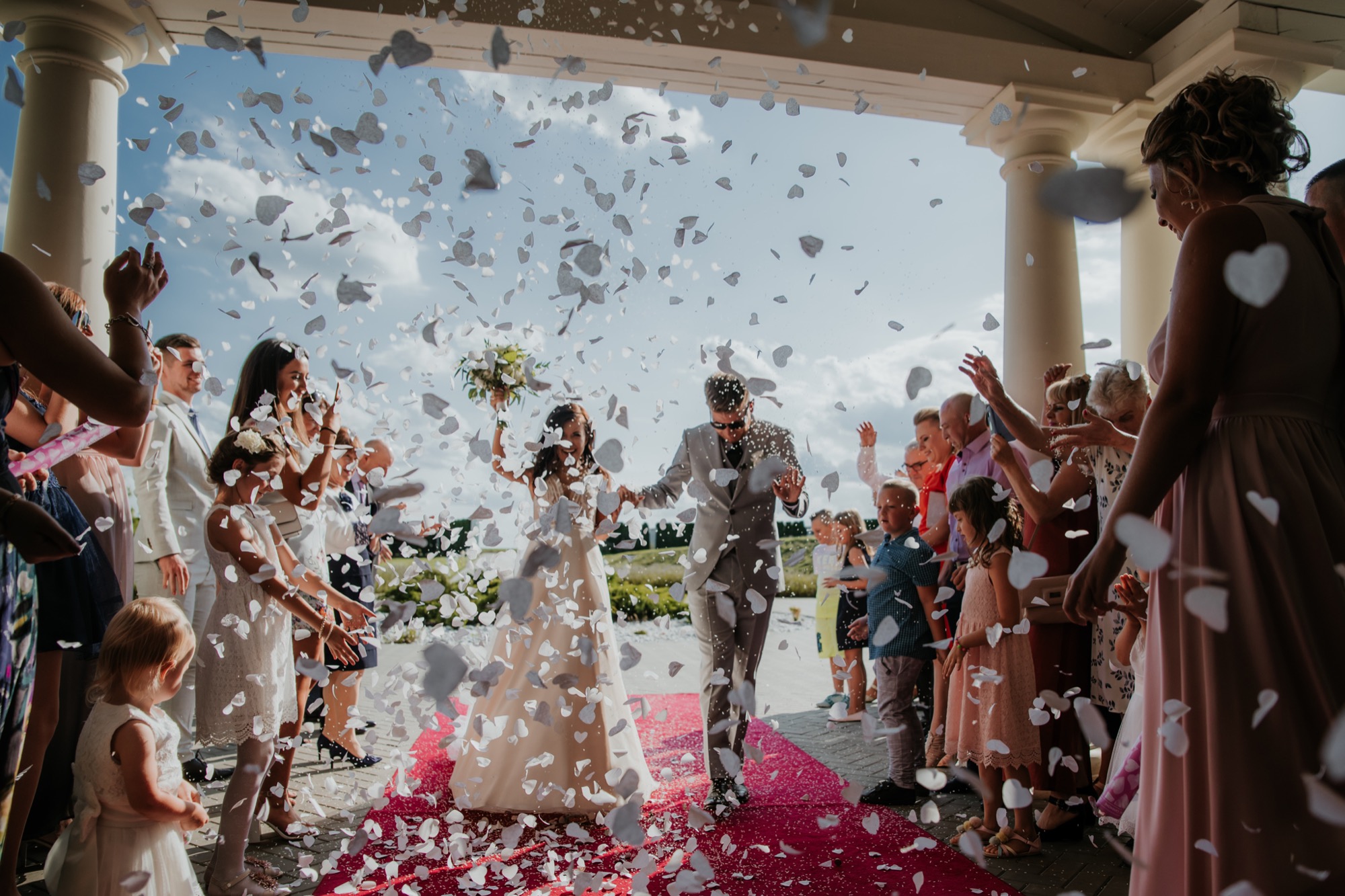 Para młoda wychodzi z ceremonii ślubnej, obsypywana konfetti w kształcie serc przez gości, na tle kolumn i błękitnego nieba.